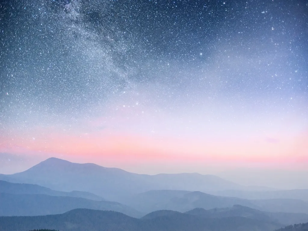 Majestic mountains undulate into the distance beneath a radiant night sky. The peaks are softly rendered in shades of blue, fading into a pale pink horizon. Above, a vast expanse of stars twinkles against the inky expanse. This peaceful image evokes tranquility and wonder, ideal for projects centered on nature, exploration, and the beauty of the night sky.